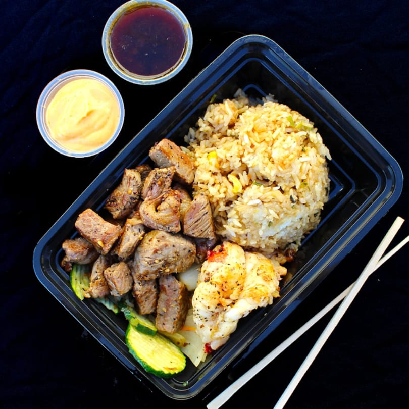 Hibachi steak and shrimp with fried rice, zucchini, and two sauces in a to-go container.